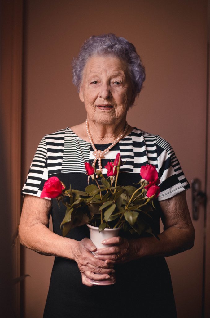 Femme âgée portant une robe noire et blanche ainsi qu'un collier de perles et qui tient un bouquet de fleur. 